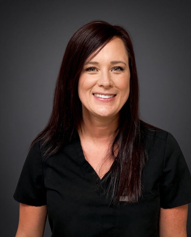 Headshot of a smiling female staff member with long dark hair wearing black scrubs against a grey backdrop