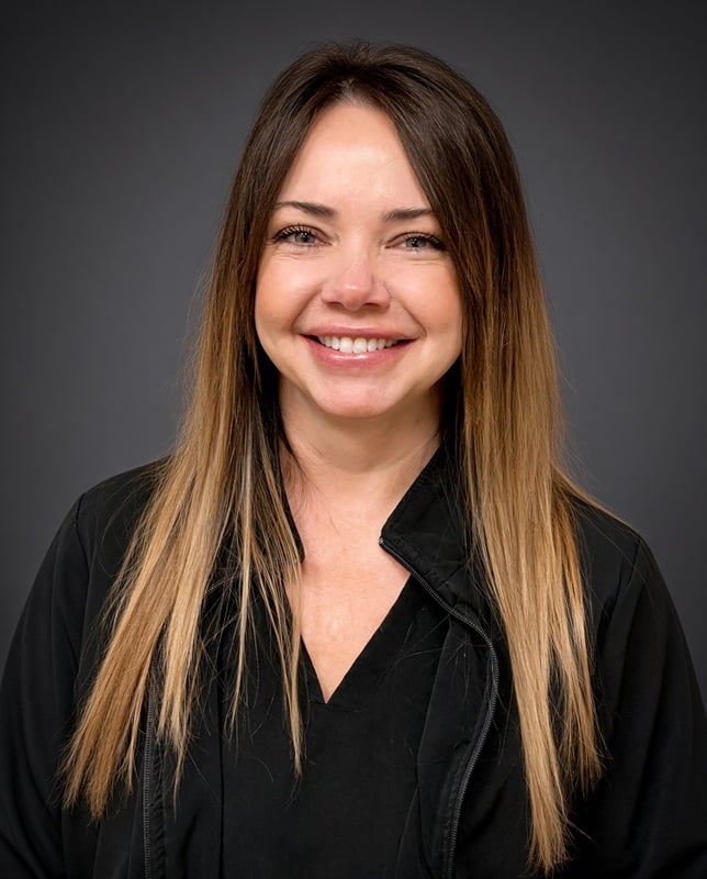 Headshot of a smiling female staff member with long highlighted hair wearing a black jacket against a studio backdrop