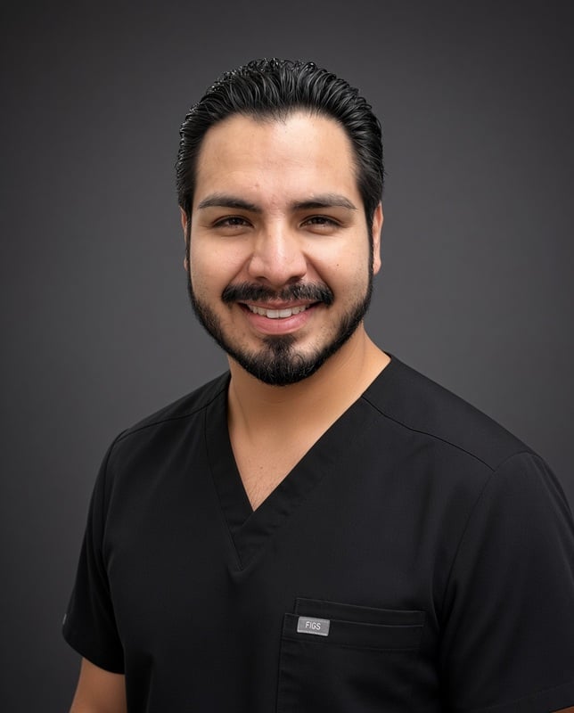Headshot of a smiling male dental staff member with dark hair and beard wearing black scrubs against a grey backdrop