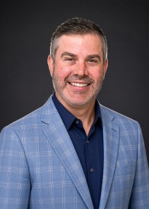 Headshot of Dr. Dean Lankford smiling in a blue plaid blazer against a dark grey background